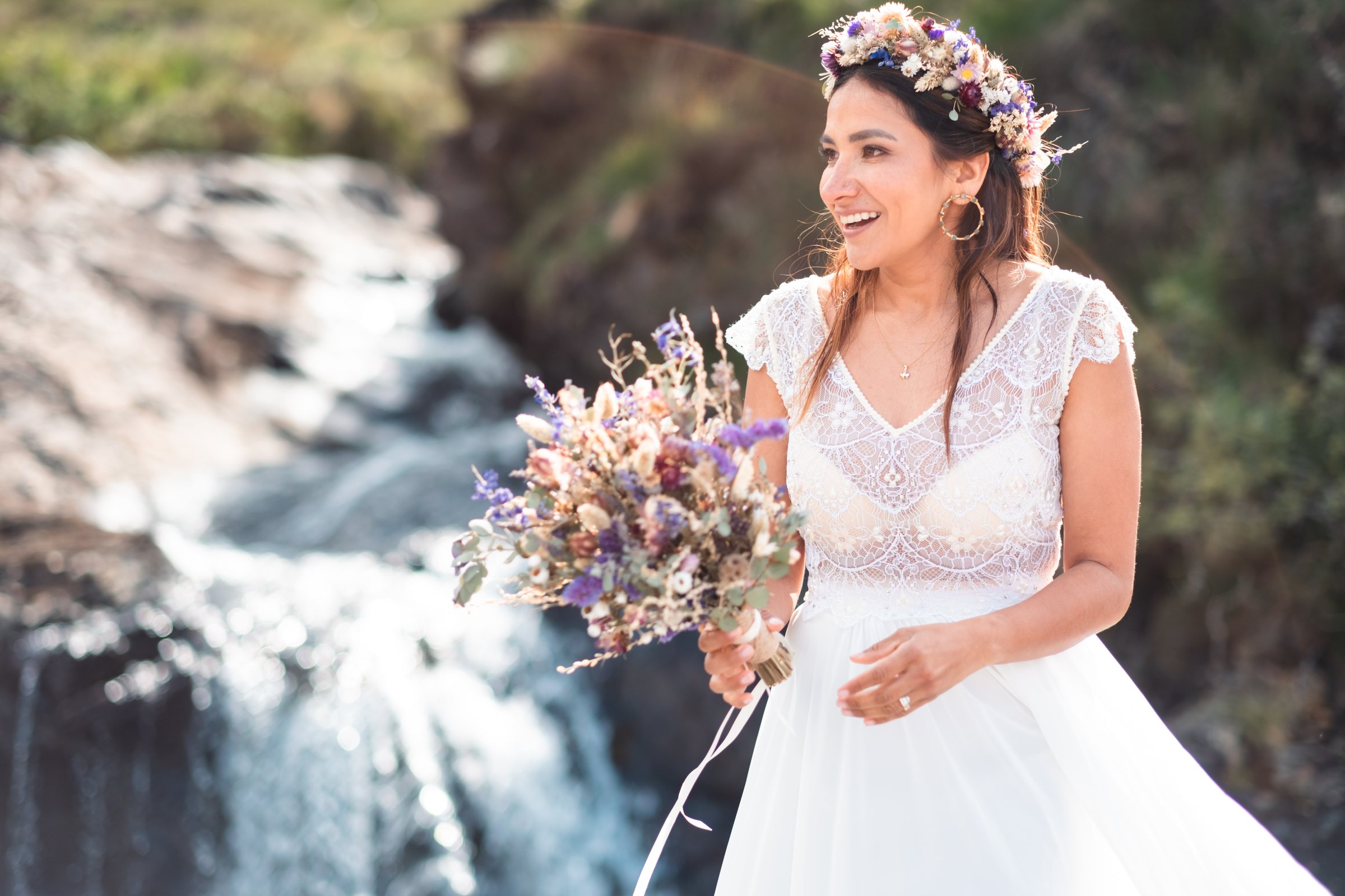A bride holding a dried flower bouquet and wearing a dried flower crown beside a waterfall